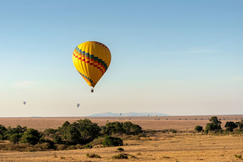 Featured in the Jaya Travel & Tours blog "Movie Locations from Out of Africa" this image shows a yellow hot air balloon flying over the trees and desert sands of Maasai Mara National Reserve in Kenya.