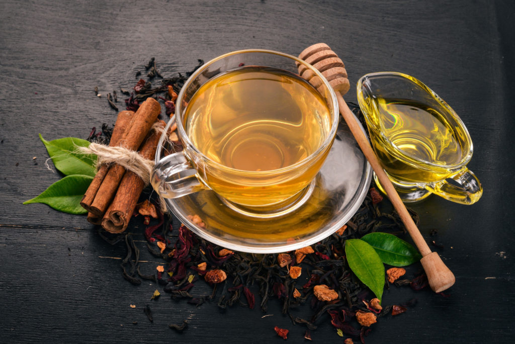 A cup of tea in a clear tea cup and saucer nestled between a pitcher of sweet honey and bound cinnamon sticks.