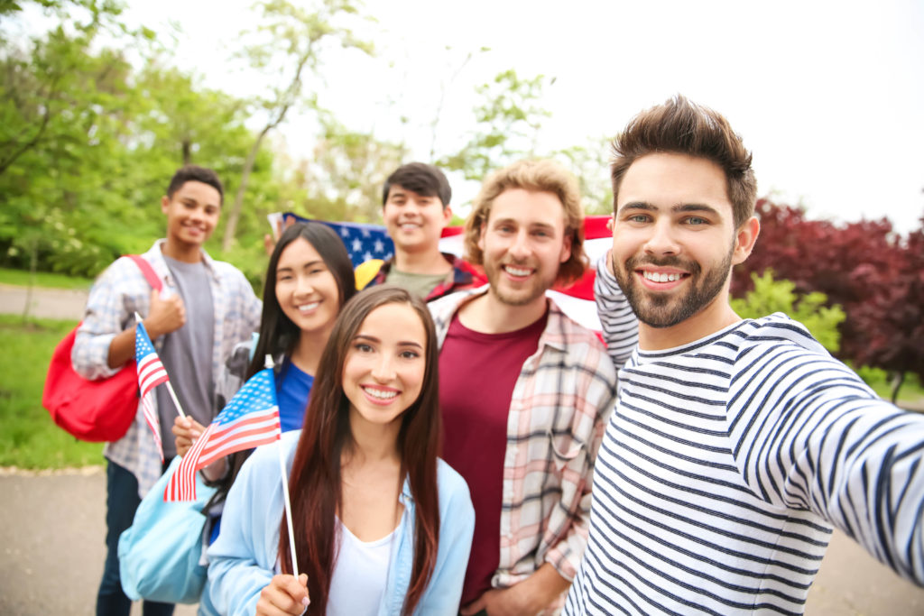 Group of American students at university on travel abroad tour taking selfie outdoors with an American flag on their study abroad tour program.