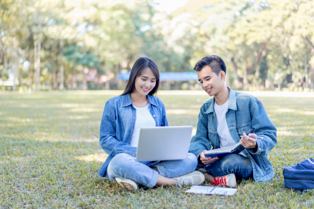 Male and female university students laying on the grass studying with a laptop and a book on their travel abroad program trip.