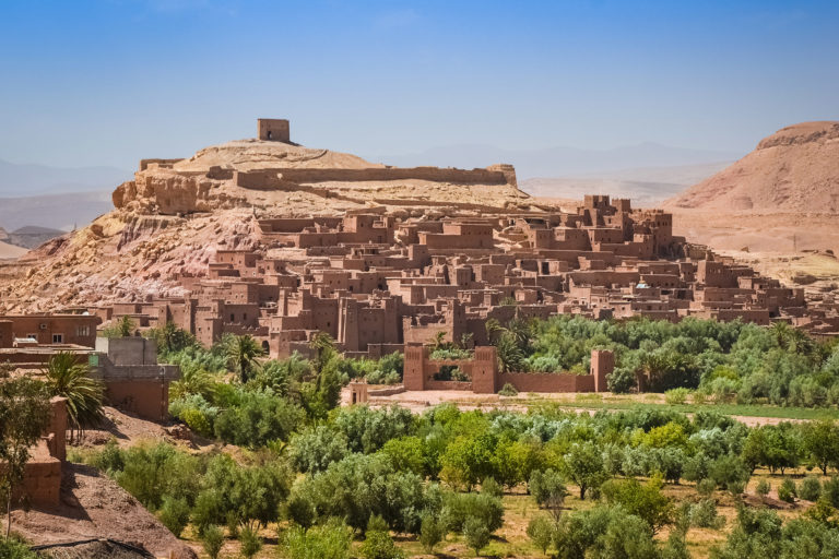 The red city, Ksar of Ait-Ben-Haddou, is located in the Province of Ouarzazate, Morocco. The Ultimate Morocco Experience customized tour by Jaya Travel & Tours visit to the outskirts of Kasbah of Aït Benhaddou in the distance with a blue sky surrounded by golden, sandy mountains.