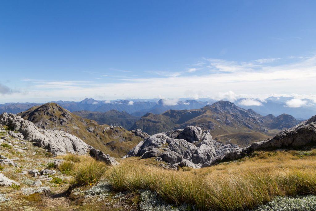 A photo of Mount Owen National Park in New Zealand, Australia, where the top of rocky and grassy mountains are at the same height as the nearby clouds, and highlighted by the bright blue sky. This photo is featured in Jaya Travel & Tours blog, "On Location: The Lord of the Rings," which describes the real life filming locations of famous movies, with photo credit from IMDB.