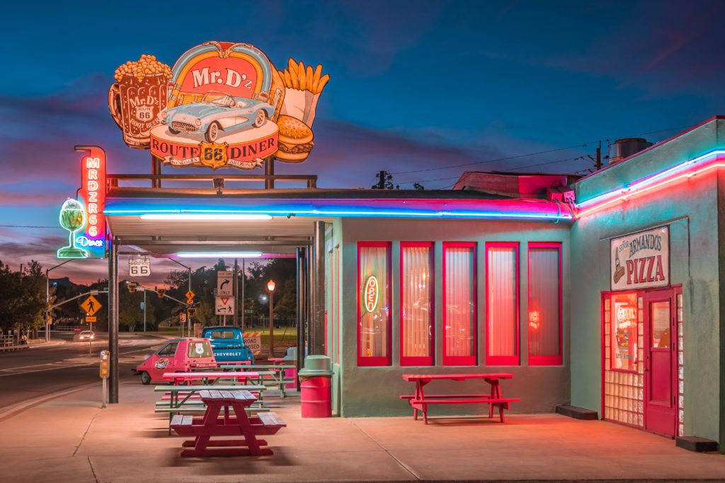 A landscape shot of a diner on Route 66 selling burgers, fries, and root beer, which is lit up by old signs and neon lights. This image is featured in the Jaya Travel & Tours blog, "A United States Road Trip," which is a travel guide to the best road trips and driving the best highways in America.