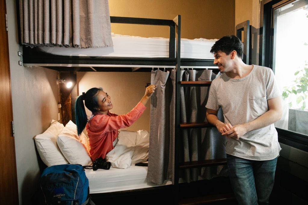 A young man and woman are in their private hostel room which includes bunk beds, privacy curtains, personal lights, storage, and more! This image is featured in the Jaya Travel & Tours blog post, "Debunking 5 Myths About Hostels," which describes false lies about staying in a hostel on vacation and why they are incorrect.