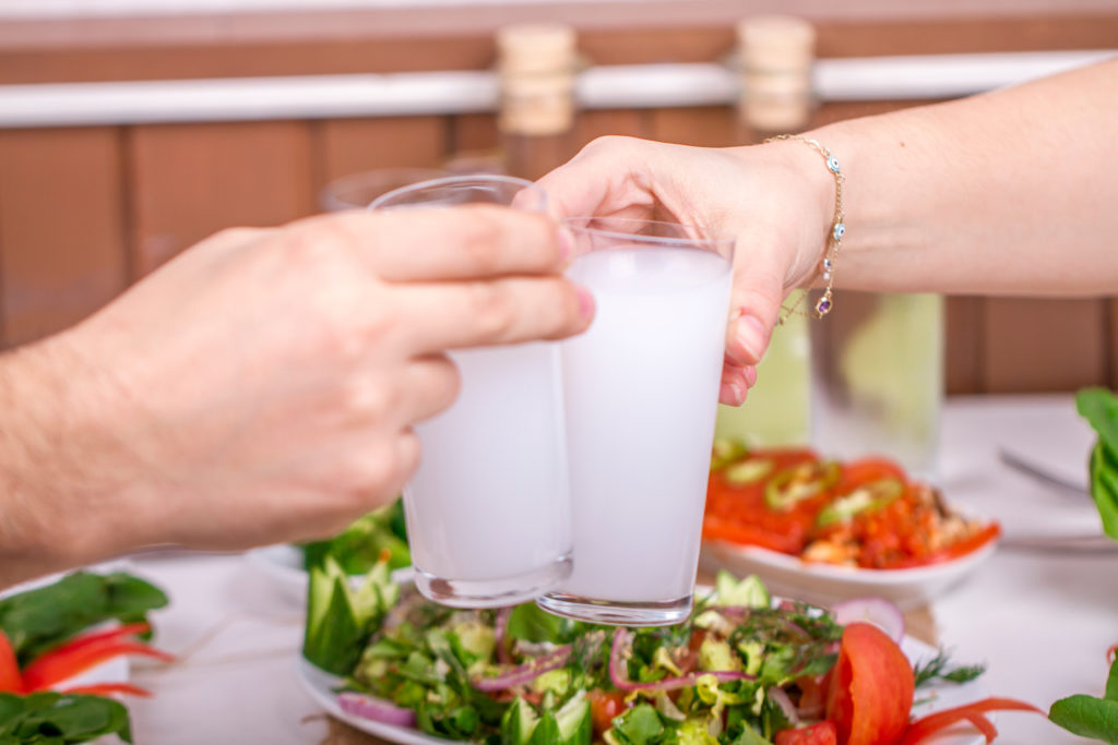 Two hands reach forward over their dinner plates with salad to clink their glasses of raki, a traditional alcoholic beverage from Turkey. This image is featured in the blog from Sky Bird Travel & Tours, "A Guide To Turkish Cuisine," which describes the types of food, drink, desserts, and dishes from Turkey.