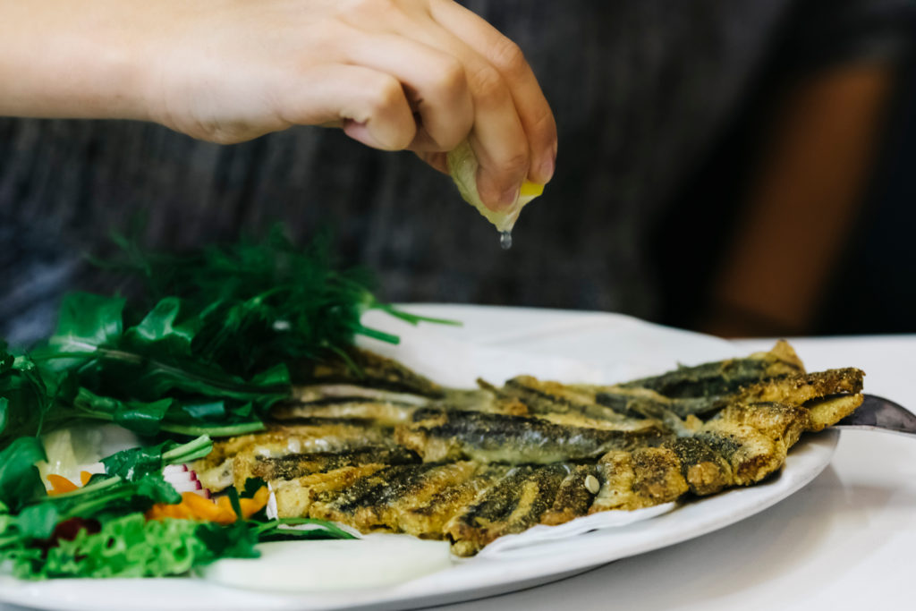 A plate of traditional hamsi, or fried sardines from the Black Sea region, is presented on a table with a hand squeezing fresh lemon over top. This image is featured in the blog from Sky Bird Travel & Tours, "A Guide To Turkish Cuisine," which describes the types of food, drink, desserts, and dishes from Turkey.