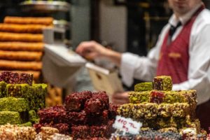 Multiple stacks of gourmet Turkish delights can be seen through the window of a sweet store, with a baker in the background. This is the featured image in the blog from Sky Bird Travel & Tours, "A Guide To Turkish Cuisine," which describes the types of food, drink, desserts, and dishes from Turkey.