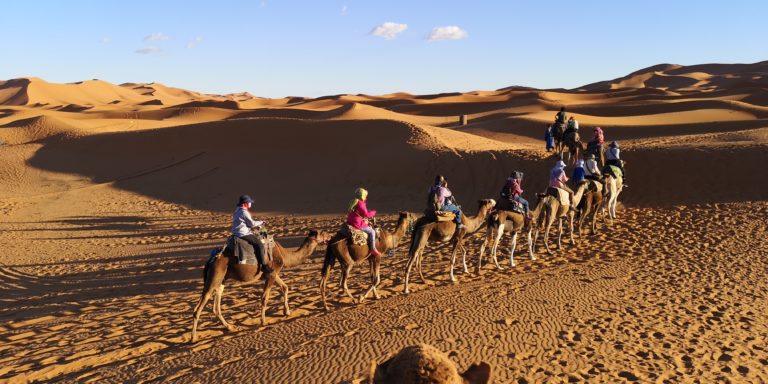 Featured in Jaya Travel & Tours blog post "Jaya's Ultimate Morocco Tour," this image features a group of tourists riding camels in the desert.